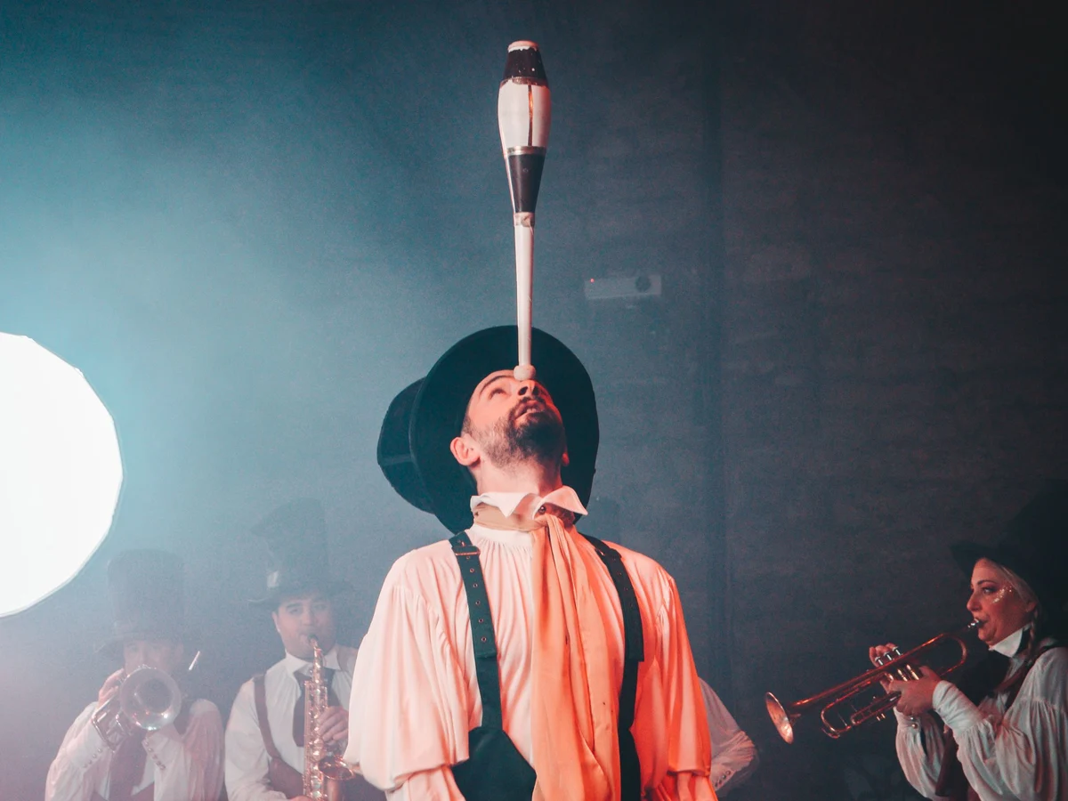 A costumed circus-style performer balancing a club on his chin while a brass band plays behind him (representative photograph, not Avanspettacolo Venezia's own cast)